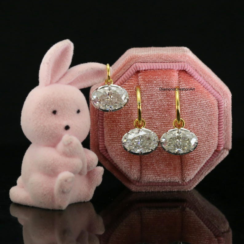 May include: A pair of oval-cut clear gemstone earrings with gold-toned hoops, displayed on a pink velvet octagonal jewellery box next to a small pink rabbit figurine. The text DiamondCreationArt is visible on the velvet surface of the box.