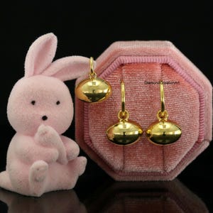May include: A pair of polished gold hoop earrings with smooth, rounded, oval-shaped charms suspended from each hoop. The earrings are displayed alongside a light pink, flocked, rabbit-shaped figurine and a pink velvet octagonal jewellery box.
