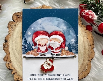 May include: A Christmas-themed friendship bracelet with a charm of two Santa Clauses. The card reads "Close your eyes and make a wish, then tie the string around your wrist. When the bracelet breaks in two, the wish you made just might come true."