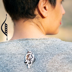 May include: A silver necklace with a pendant featuring intricate Arabic calligraphy. The pendant is made of a dark brown material and hangs from a thin silver chain.