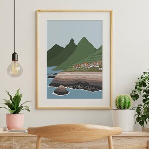 May include: A framed illustration of a coastal landscape with a small village nestled in the hills. The scene features a blue ocean, green hills, and a rocky coastline.