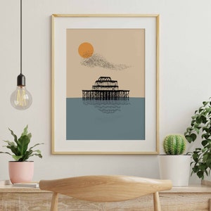 May include: A framed print of a black and white illustration of a pier with a large sun and a flock of birds in the sky. The background is a light brown color with a blue ocean.
