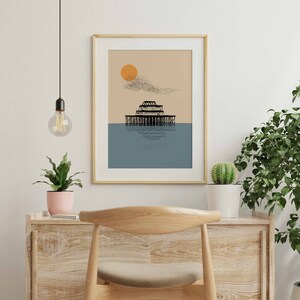 May include: A framed print of a black and white illustration of a pier with a large orange sun in the background. The pier is reflected in the blue water below.