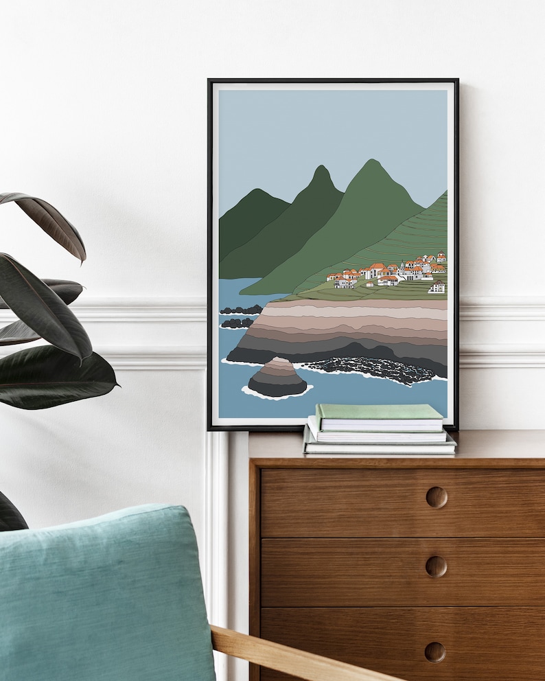 May include: A minimalist line art print of a coastal landscape with green mountains, a small village, and a rocky shoreline. The print is framed in black and has a light blue background.