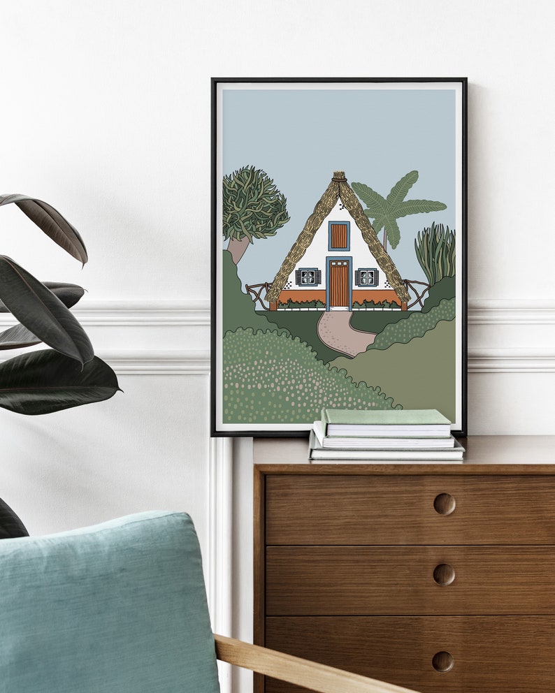 May include: A framed illustration of a white cottage with a thatched roof, surrounded by green foliage and palm trees. The cottage has a brown door and two windows with blue frames.