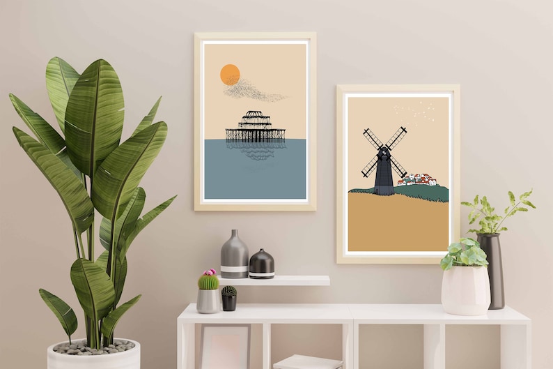 May include: Two framed illustrations, one depicting a pier with a sun and birds in the sky, the other depicting a windmill with a small village in the background. Both illustrations are in a minimalist style with muted colors.