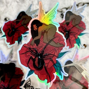 May include: Holographic stickers featuring a fairy with iridescent wings, perched on a red rose with a black spider. The fairy has long black hair and a nude-colored body. The stickers have a glossy finish.
