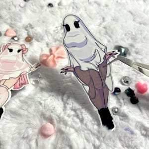 May include: A white ghost sticker featuring a female figure wearing a white sheet and black fishnet stockings. The sticker is on a white fluffy surface.
