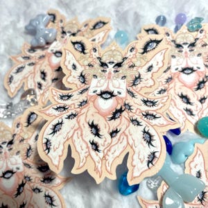 May include: A collection of stickers featuring a whimsical, multi-eyed creature with a peach and cream color palette. The creature has multiple eyes, eyelashes, and a detailed design. The stickers are surrounded by colorful glass beads.