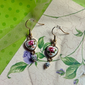 May include: A pair of gold heart-shaped earrings with a floral design and a clear crystal dangle. The earrings are on a white background with green leaves.