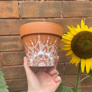 May include: A terracotta plant pot featuring a white mandala pattern. The pot is held up, with a brick wall and a sunflower in the background. The mandala design is hand-painted, adding a decorative element to the pot.