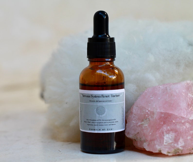 May include: A brown glass bottle with a black dropper top and a white label that reads "Nervous System Reset Tincture". The bottle is sitting on a white surface next to a large pink crystal.