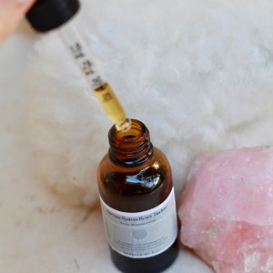 May include: A brown glass bottle with a dropper filled with a clear liquid. The bottle is labeled "Nervous System Reset Tincture". A pink crystal is next to the bottle.