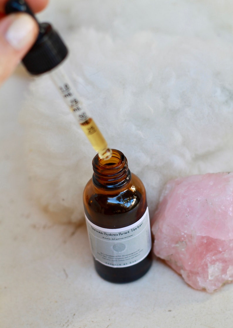 May include: A brown glass bottle with a dropper filled with a clear liquid. The bottle is labeled "Nervous System Reset Tincture". A pink crystal is next to the bottle.