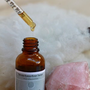 May include: A brown glass bottle with a dropper filled with a clear liquid. The bottle is labeled "Nervous System Reset Tincture -  Botanical Support for the Autonomic Nervous System". The bottle is sitting on a white fluffy surface next to a large pink crystal.