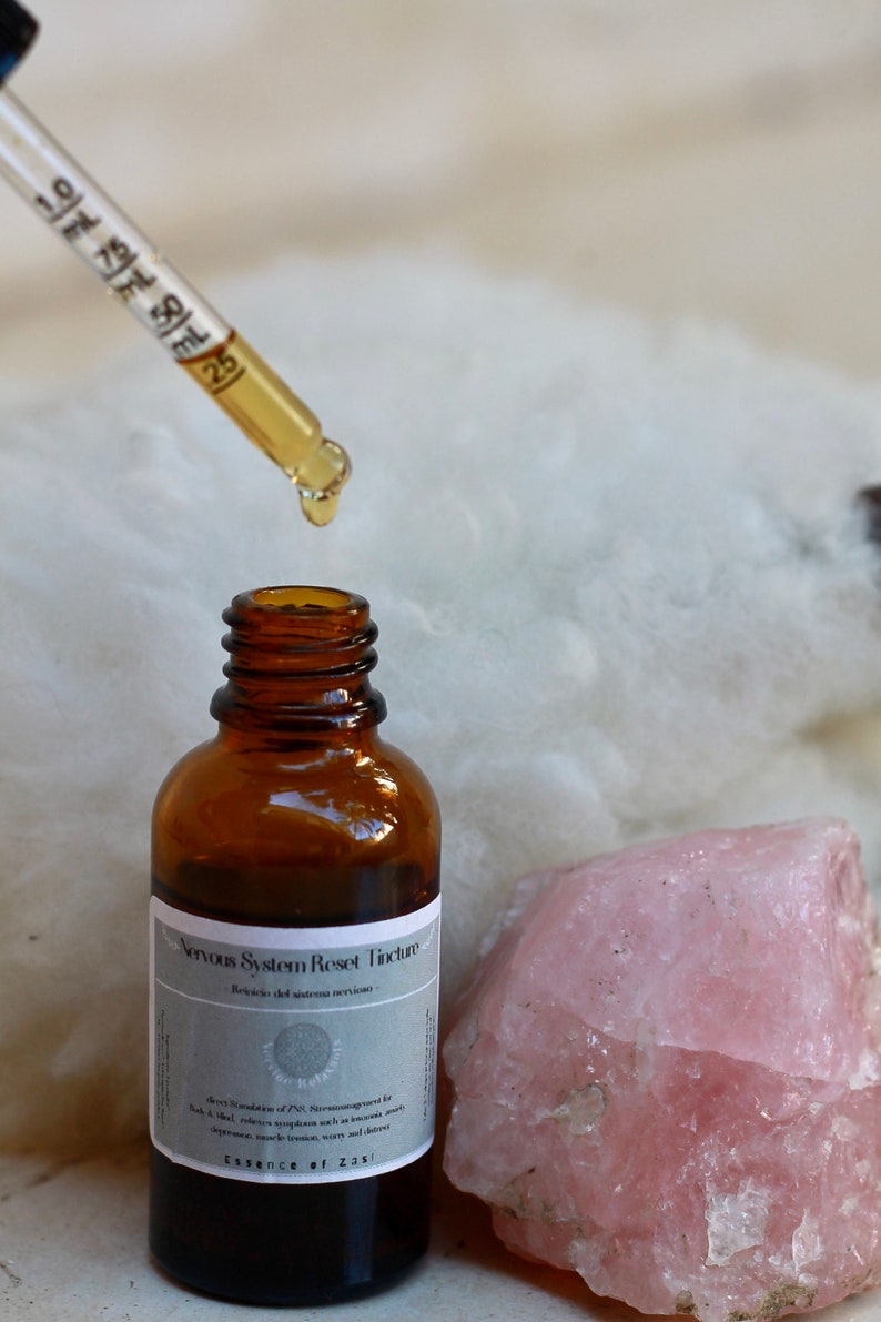 May include: A brown glass bottle with a dropper filled with a clear liquid. The bottle is labeled "Nervous System Reset Tincture -  Botanical Support for the Autonomic Nervous System". The bottle is sitting on a white fluffy surface next to a large pink crystal.