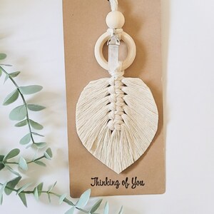 Peut inclure: Une feuille en macramé blanche avec un cristal suspendu à un anneau en bois. L'arrière-plan est brun avec le texte "Thinking of You".