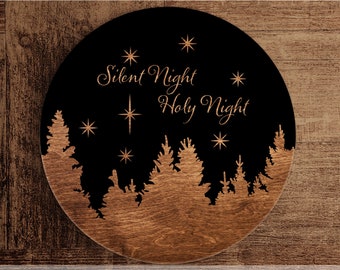 May include: A round wooden sign with a black background and a white silhouette of a forest with stars. The text "Silent Night Holy Night" is written in a cursive font in the center of the sign.
