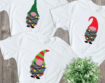 Könnte beinhalten: Drei weiße T-Shirts mit einem Weihnachtszwerg-Design. Jeder Zwerg trägt einen Hut in einer anderen Farbe: grün, rot und rot-grün gestreift. Jeder Zwerg ist in Lichterketten mit bunten Glühbirnen gewickelt.