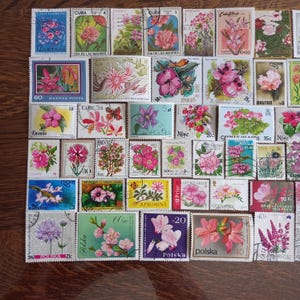 Può includere: Una collezione di francobolli vintage con disegni floreali colorati. I francobolli presentano vari fiori in tonalità di rosa, viola e rosso, con testo in più lingue. I francobolli sono disposti su una superficie di legno.