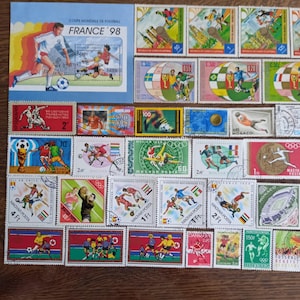 Può includere: Una collezione di francobolli colorati con giocatori di calcio in azione. I francobolli presentano vari disegni, tra cui il francobollo "FRANCE '98", e francobolli provenienti da Ungheria, Monaco e altri paesi. I francobolli sono disposti su una superficie di legno.