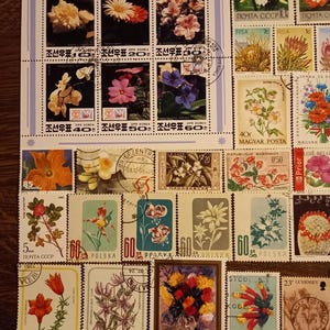 Op de afbeelding: Een verzameling kleurrijke postzegels met bloemmotieven. De postzegels tonen verschillende bloemen, waaronder orchideeën, madeliefjes en tulpen, met tekst in meerdere talen. De postzegels zijn gerangschikt op een lichtgekleurde achtergrond.