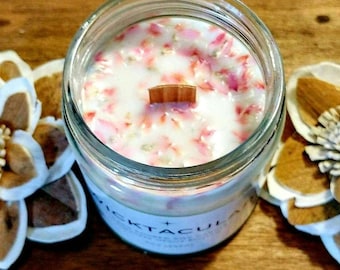 May include: A clear glass jar candle with a wooden wick, filled with white wax and pink flower petals. The candle is surrounded by decorative dried flowers on a wooden surface. The candle has a label with text.