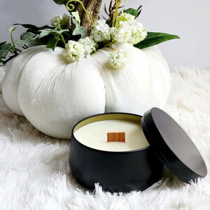 May include: A black candle in a metal tin with a wooden wick. The candle is sitting on a white fluffy surface next to a white pumpkin.