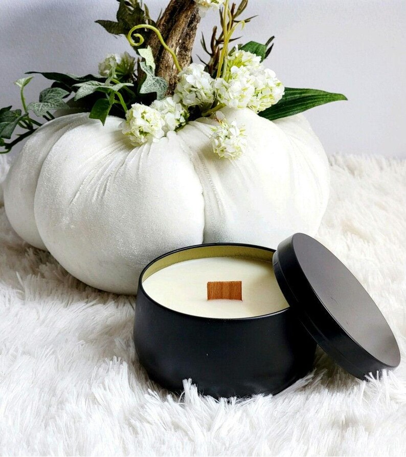 May include: A black candle in a metal tin with a wooden wick. The candle is sitting on a white fluffy surface next to a white pumpkin.