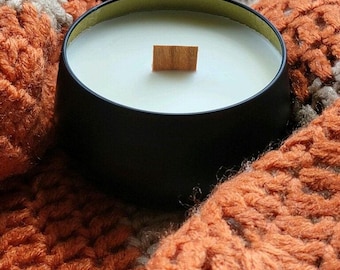 May include: A black candle with a wooden wick in a black and gold tin container. The candle is sitting on a knitted orange and beige blanket.