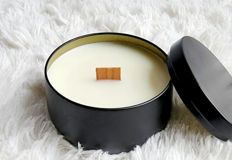 May include: A black candle jar with a wooden wick and a white wax candle inside. The jar has a gold rim and is sitting on a white fluffy surface.
