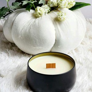 May include: A black candle with a wooden wick in a black metal jar. The candle is sitting on a white fluffy surface. A white velvet pumpkin is in the background.