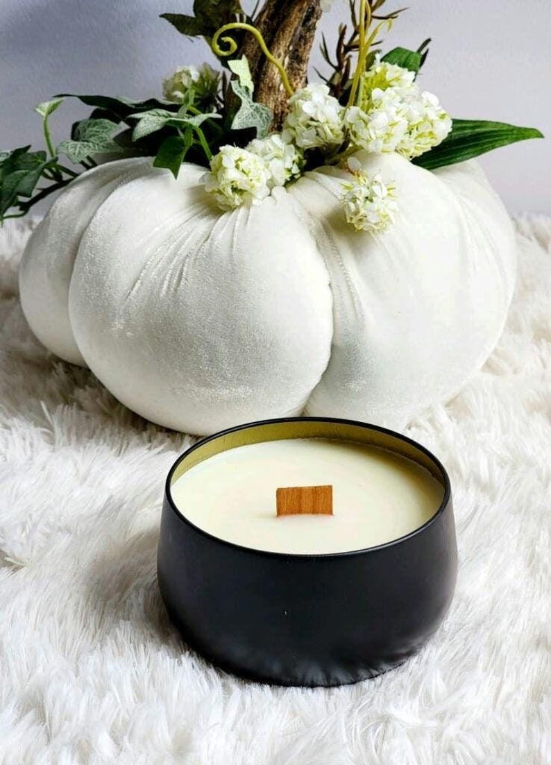 May include: A black candle with a wooden wick in a black metal jar. The candle is sitting on a white fluffy surface. A white velvet pumpkin is in the background.