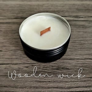 May include: A black candle tin with a white wax interior and a wooden wick. The text "Wooden wick" is written in a cursive font at the bottom of the image. The candle sits on a wooden surface.