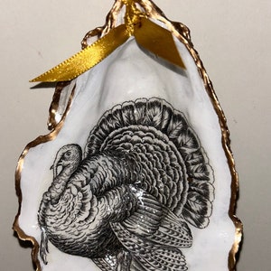 May include: A white oyster shell ornament with a gold trim, featuring a black and white illustration of a turkey. The ornament has a gold ribbon and a gold wire hanger.