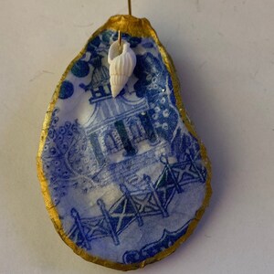May include: A blue and white oyster shell with a gold rim, featuring a printed design of a house and fence. A small white seashell is attached to the top of the shell.