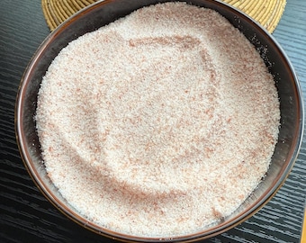 May include: A dark brown bowl filled with pink Himalayan salt. The salt has a fine, granular texture and is a light pink color. A woven straw hat is in the background.
