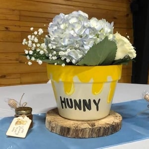 May include: A yellow ceramic flower pot with the word "Hunny" written on it. The pot is decorated with yellow drips and holds a bouquet of white hydrangeas and baby's breath. The pot is sitting on a wooden slice.