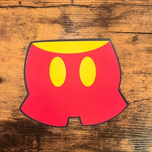 Puede incluir: Un recorte de papel rojo de pantalones cortos de personaje de dibujos animados con dos óvalos amarillos y una cintura amarilla. Los pantalones cortos tienen un contorno azul oscuro y están sobre un fondo de madera.