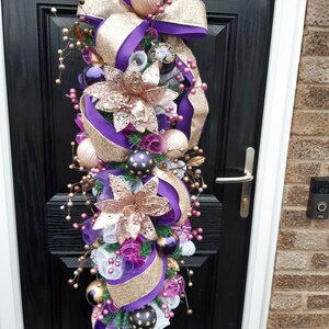 May include: A purple and gold Christmas swag with a large bow, baubles, and floral accents. The swag is hanging on a black door.