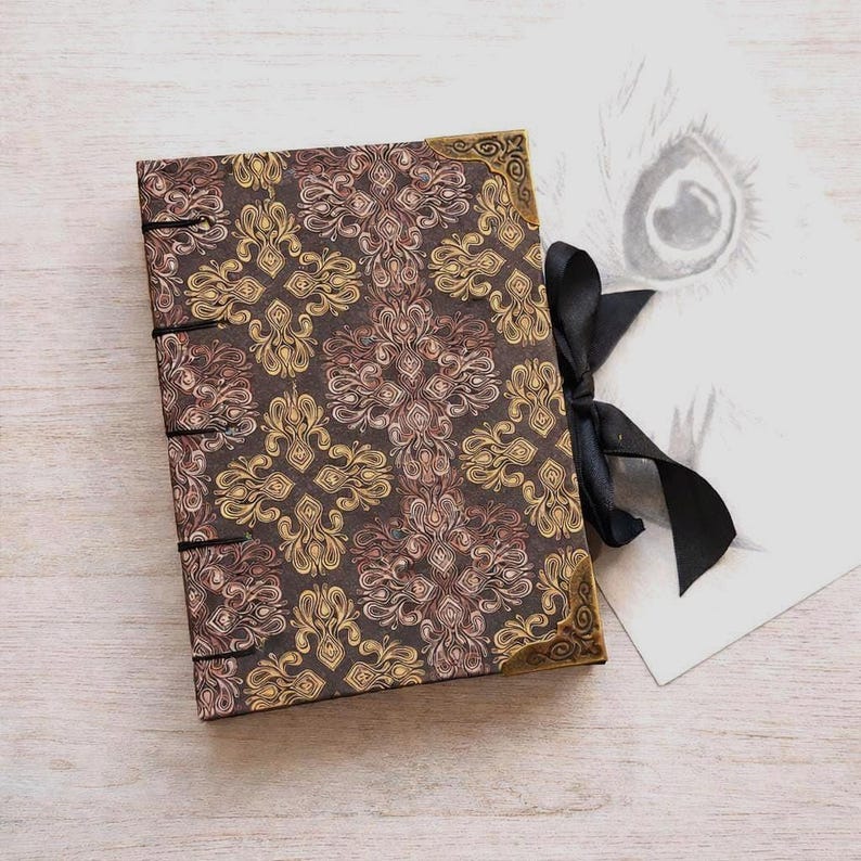 Puede incluir: Un peque&ntilde;o diario decorativo con una cubierta marr&oacute;n que presenta un patr&oacute;n floral repetido en oro y rosa. El diario tiene protectores de esquina dorados, encuadernaci&oacute;n negra y un cierre de cinta negra. Un dibujo de un ojo es visible en el fondo.