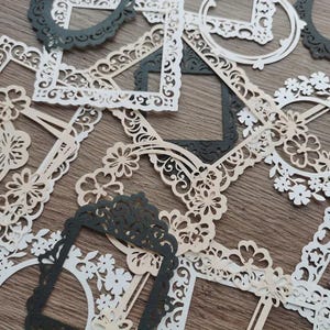 Può includere: Una collezione di cornici di carta tagliate in modo intricato in varie forme e dimensioni. Le cornici sono in tonalità di bianco, crema e grigio scuro, con disegni floreali e ornati. Alcune cornici sono quadrate, altre rotonde e alcune hanno bordi smerlati.