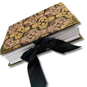 Puede incluir: Un diario decorativo con una cubierta con estampado floral negro, dorado y marr&oacute;n. El diario est&aacute; encuadernado con una cinta de sat&eacute;n negro atada con un lazo. Los bordes de las p&aacute;ginas son dorados.