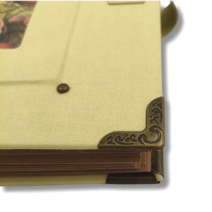 May include: Close-up of a vintage photo album with a light yellow linen cover. The album features decorative bronze corner protectors and a small, dark brown button. The pages are visible, and a green ribbon is attached.