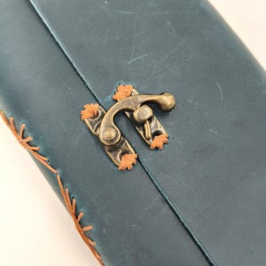 Puede incluir: Primer plano de un diario de cuero verde azulado con un cierre de lat&oacute;n. El diario presenta costuras naranjas a lo largo de los bordes y alrededor del cierre. El cierre es de estilo vintage, lo que se suma a la est&eacute;tica artesanal.