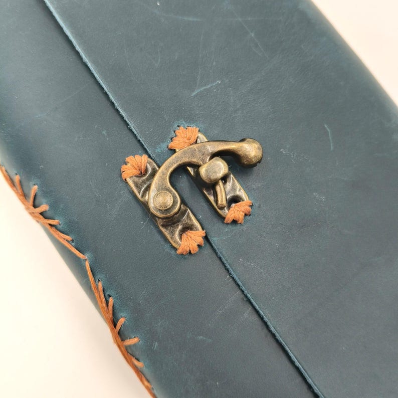 Puede incluir: Primer plano de un diario de cuero verde azulado con un cierre de lat&oacute;n. El diario presenta costuras naranjas a lo largo de los bordes y alrededor del cierre. El cierre es de estilo vintage, lo que se suma a la est&eacute;tica artesanal.