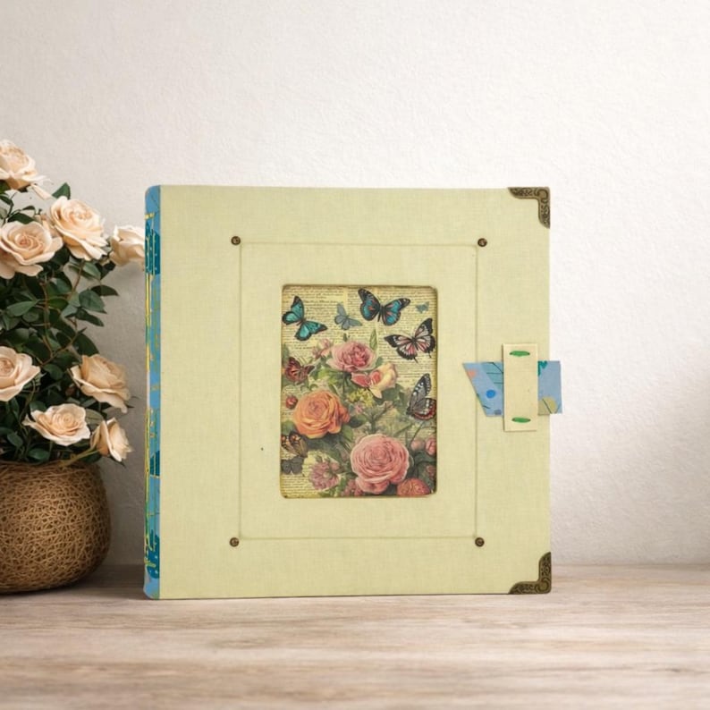 May include: A square, light yellow photo album with a floral and butterfly design. The album features a rectangular window showcasing roses and butterflies. The corners are adorned with bronze-colored metal accents, and a blue and white patterned clasp secures the album.