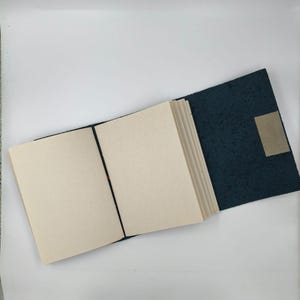 Puede incluir: Un diario abierto con p&aacute;ginas en blanco y una cubierta azul oscuro texturizada. Una banda el&aacute;stica negra mantiene el diario abierto. Una peque&ntilde;a etiqueta rectangular de color beige est&aacute; adherida a la contraportada.