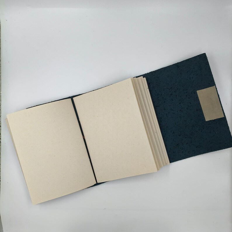 Puede incluir: Un diario abierto con p&aacute;ginas en blanco y una cubierta azul oscuro texturizada. Una banda el&aacute;stica negra mantiene el diario abierto. Una peque&ntilde;a etiqueta rectangular de color beige est&aacute; adherida a la contraportada.