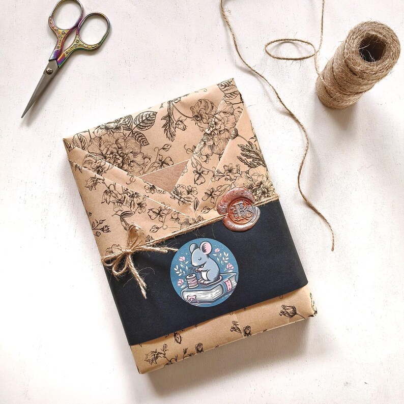 Puede incluir: Un paquete envuelto en papel floral, con una banda negra y una pegatina de rat&oacute;n. El paquete est&aacute; atado con hilo y sellado con un sello de cera. Tambi&eacute;n se ven tijeras y un carrete de hilo.